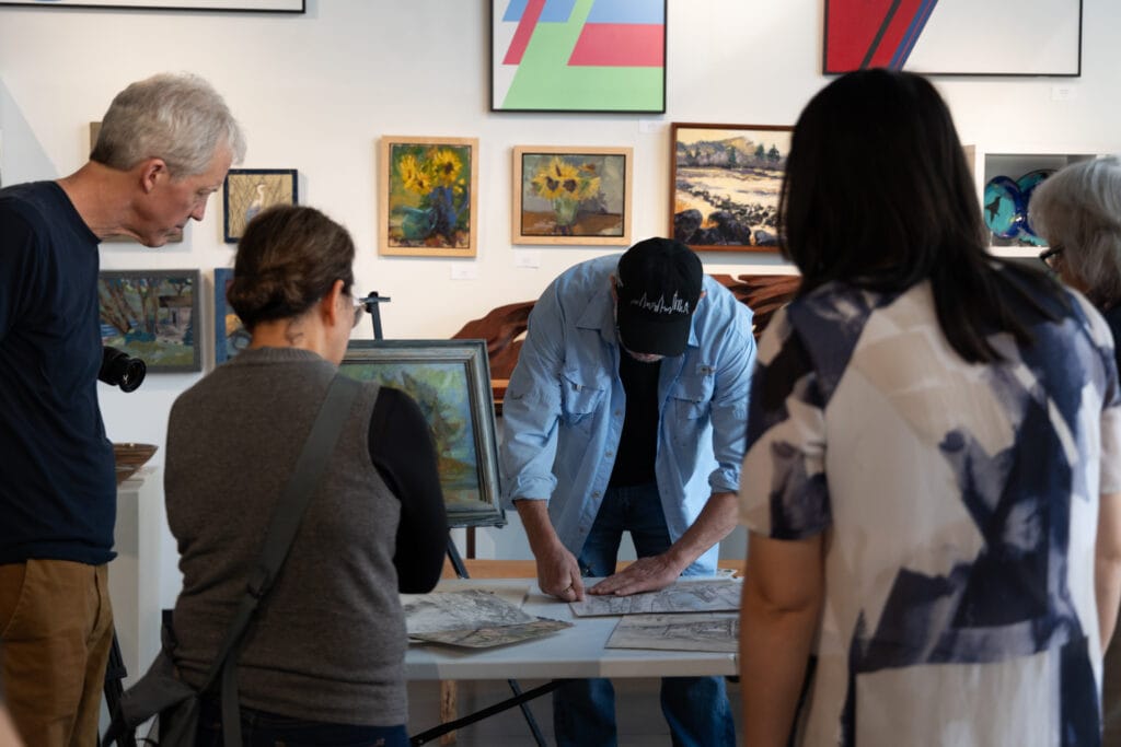 Gallery visitors gather around a table where Oregon artist Erik Sandgren demonstrates his watercolor painting process. Taken October 19th, 2024.